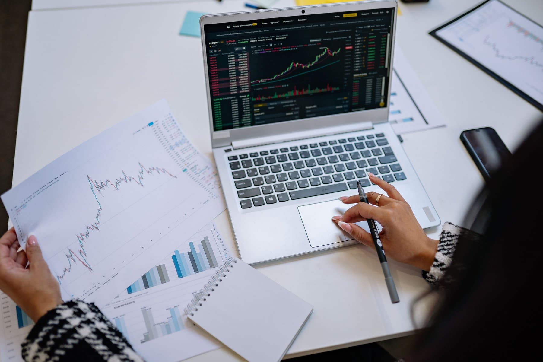 A laptop showing financial charts next to a hand holding a pen over a printed graph