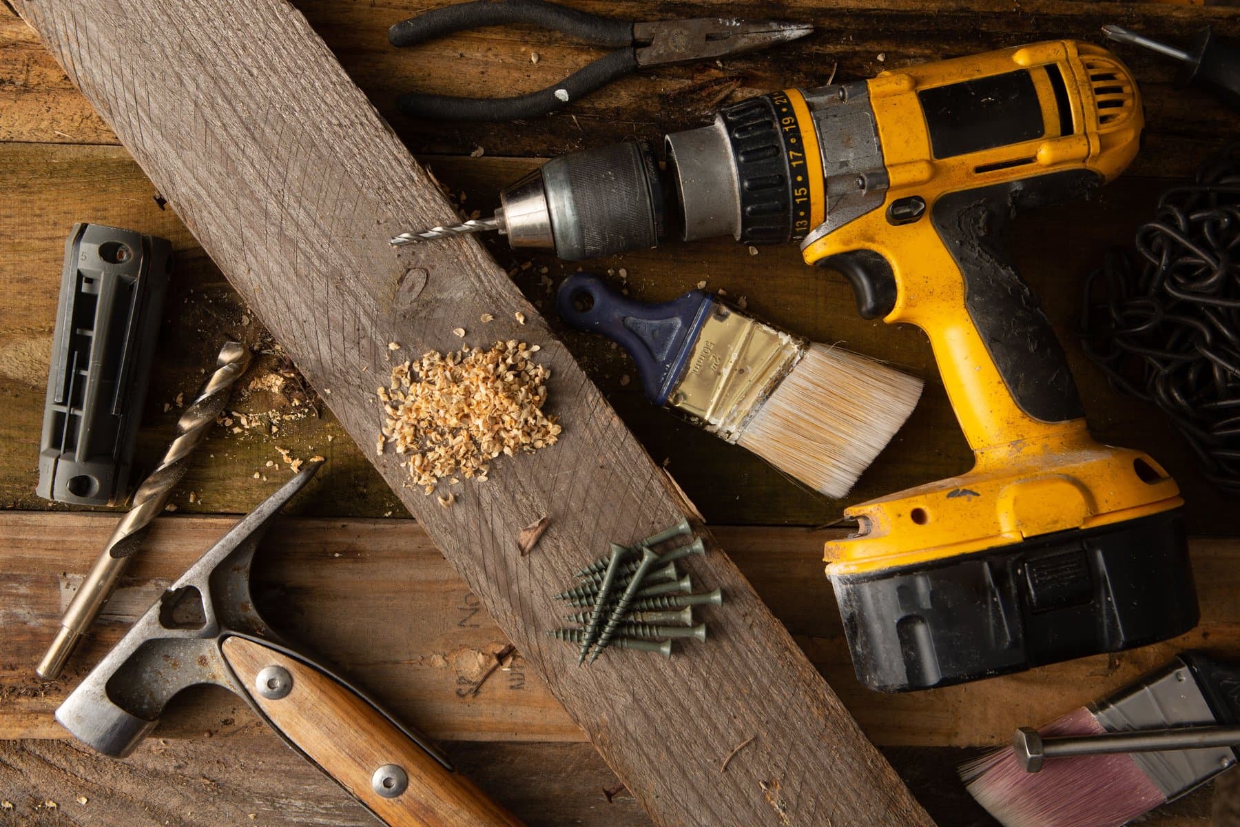 A workbench flat-lay with a drill, hammer, paintbrush, and screws